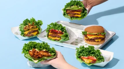 Assorted Shake Shack burgers, including lettuce-wrapped and bun options, displayed on a tray and held by hands against a light blue background.