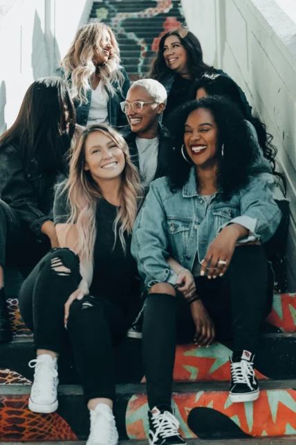 A diverse group of seven women sit together on colorful painted stairs, laughing and smiling, dressed casually in denim jackets, T-shirts and sneakers.