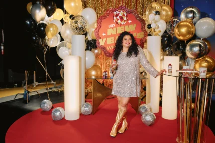 Michelle Buteau is seen in a silver sequined dress and gold boots holds a Premier Protein shake while posing on a festive New Year–themed set with balloons and branded signage.