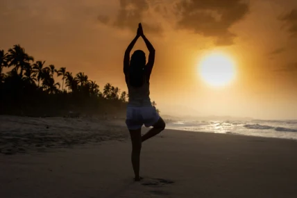 A silhouetted person balances in tree pose on a beach at sunset, with palms in prayer overhead as the sun sets over the ocean.