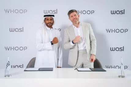 Two business executives stand side by side behind a table, each wearing a Whoop device and posing in front of a co-branded Whoop and Wasl backdrop during a signing event.